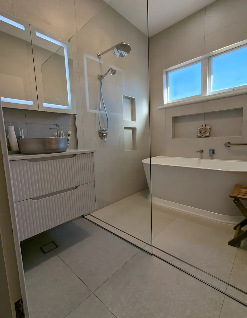Bathroom by Sunshower: white fluted vanity under a backlit mirror cabinet, a walk-in shower with two tiled niches and a freestanding bath under the window