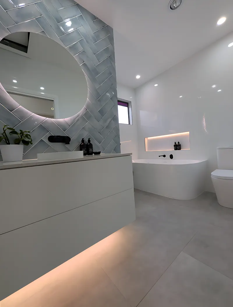 Sage green herringbone feature wall behind a round backlit mirror, with a floating vanity and a bath with a lit niche, tiled by Sunshower