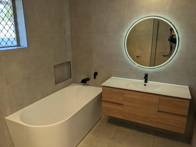 Valentine bathroom: back-to-wall bath under a tiled niche, a timber vanity and a backlit round mirror